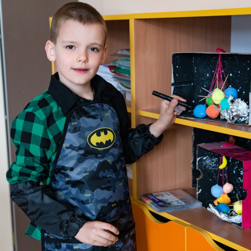 Chłopiec w fartuszku plastycznym Batman przy półce, szkolny fartuszek ochronny do malowania dla dzieci
